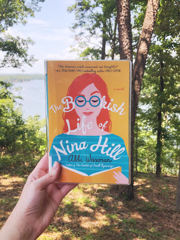 A hand holds The Bookish Life of Nina Hill by Abbi Waxman in front of a lake